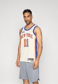 Basketball-Trikot in Creme, Blau und Orange mit dem Schriftzug "NEW YORK" und der Nummer 11 in fettem Druck. Getragen mit grauen Shorts, leichter Stoff.