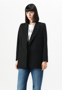 Zwarte dubbelknoop blazer van gestructureerde stof, met spitse revers, twee voorzakken en lange mouwen, gecombineerd met een wit graphic t-shirt.