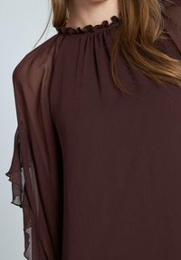 Bordeauxfarbige Bluse mit gerafftem Ausschnitt, transparenten Glockenärmeln und einem fließenden, leichten Stoff. Strukturierte Optik mit sanften Faltendetails.
