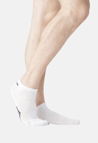 Jambes nues avec des pieds portant des chaussettes blanches à la cheville, un pied levé sur les orteils, sur un fond clair uni.