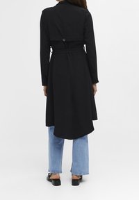 Trench-coat noir avec une taille ceinturée, des manches longues et un ourlet asymétrique. Tissu lisse avec une finition polie, orné d'un détail de bouton au dos.