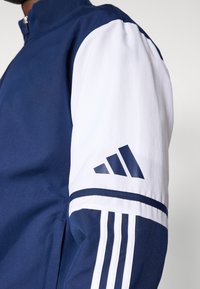 Giacca blu navy e bianca con collo con zip, caratterizzata da tre strisce bianche sulla manica e un logo adidas blu navy. Texture liscia.