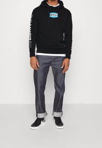 Hombre con sudadera negra con el logo "Replay", jeans oscuros con dobladillo y zapatillas negras de Nike, de pie contra un fondo liso.