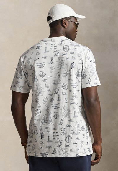 Άντρας με λευκό καπέλο και λευκό T-shirt με μαύρα ναυτικά σχέδια, όπως άγκυρες, πλοία και πυξίδες, γυρισμένος ανάποδα.