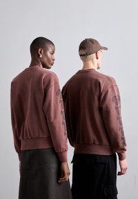 Brun sweatshirt lavet af bomuld, med ribbede manchetter og kant, med grafisk tekst trykt på ærmerne. Båret af to figurer set bagfra.