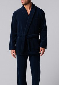 Uomo che indossa un accappatoio in velluto blu navy con colletto a scialle, cintura legata, tasche frontali e sul petto, e pantaloni abbinati su uno sfondo semplice.