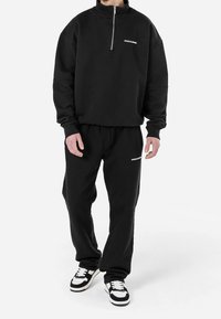 Felpa nera con zip e colletto alto, abbinata a pantaloni della tuta neri coordinati. Entrambi presentano dettagli del logo in bianco.