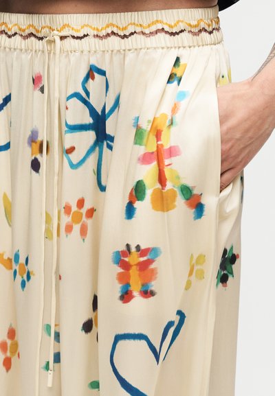 Χέρι με καπέλο ξεκουράζεται στην τσέπη κρεμ παντελονιού με πολύχρωμα αφηρημένα λουλουδένια σχέδια και ελαστική μέση με κορδόνι.