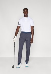Polo blanco con cuello y rayas en las mangas, combinado con pantalones gris oscuro y zapatos blancos de golf. Sosteniendo un palo de golf.