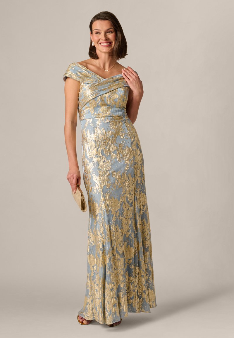 Frau in einem blau-goldenen, schulterfreien Kleid mit Blumenmuster, die eine beige Clutch hält, lächelnd, vor einem schlichten neutralen Hintergrund stehend.