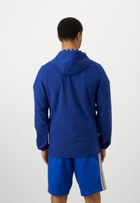 Plava vodootporna jakna s kapuljačom, dugim rukavima i aerodinamičnim dizajnom, u kombinaciji s plavim šortsima s zlatnim prugama.