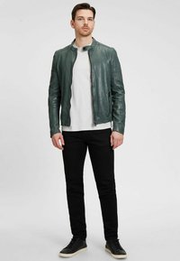 Groene leren motorjack met een hoge kraag. Voorzien van een ritssluiting, zijzakken en textuuraccenten op de schouders. Zwarte jeans en sneakers.
