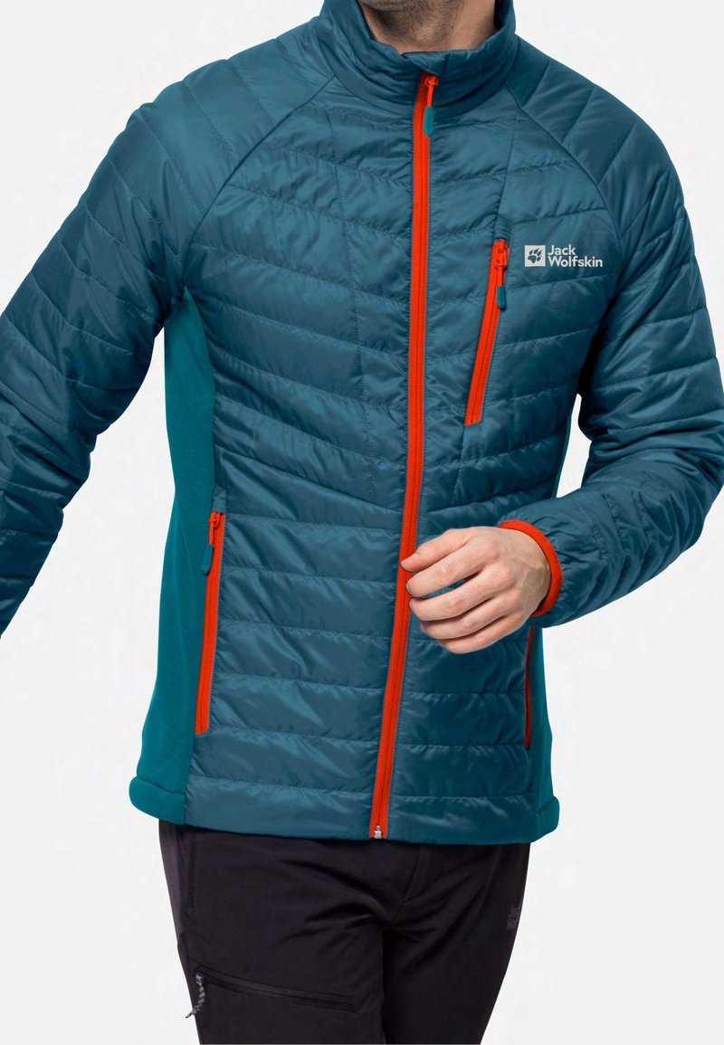 Homme portant une veste matelassée bleu sarcelle avec des fermetures éclair orange et le logo Jack Wolfskin sur la poitrine, associée à un pantalon noir.