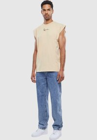 Beige sleeveless t-shirt with a Black logo, paired with light blue denim jeans and white sneakers, showcasing a relaxed fit and casual style.