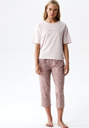 Młoda kobieta ubrana w jasnoróżową koszulkę z krótkim rękawem z napisem oraz krótkie fioletowe spodnie z białymi abstrakcyjnymi liniami, stojąca boso na białym tle.