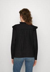Blusa nera a maniche lunghe con dettagli arricciati sulle spalle, pieghe sulla schiena e un tessuto morbido e testurizzato. Abbinata a jeans azzurri chiari.
