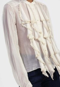 Blusa bianca trasparente con dettagli in pizzo arricciato sul davanti, chiusura a bottoni e maniche lunghe, abbinata a jeans in denim scuro.