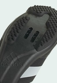 Semelle de chaussure de sport noire avec un grip en caoutchouc texturé, comportant deux évents, des picots surélevés et un logo en relief sur le côté.
