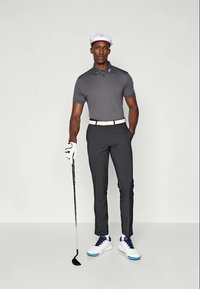 Férfi szürke pólóban, fekete nadrágban, fehér övvel, fehér golfkesztyűben, fehér sapkában és fehér golfcipőben, aki golfütőt tart egy sima háttér előtt.