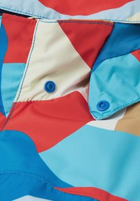 Bunt gemusterter Stoff mit großen geometrischen Formen in Rot, Blau, Creme und Braun, verziert mit blauen Druckknöpfen und kontrastierenden Nähten. Glatte Textur.
