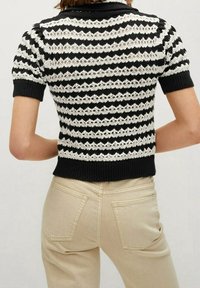 Femme portant un haut en maille à manches courtes à rayures noires et blanches avec un motif ajouré, associé à un pantalon beige taille haute, vue de dos.