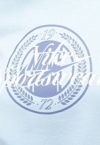 Ανοιχτό μπλε ύφασμα με κυκλικό λογότυπο Nike και την ένδειξη "Nike Sportswear" σε λευκό. Περιλαμβάνει τους αριθμούς "19" και "72."