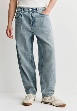 Mann trägt hellblaue locker sitzende Jeans, weißes Hemd eingesteckt und beige Sneakers mit braunen Sohlen, steht mit einer Hand in der Tasche.