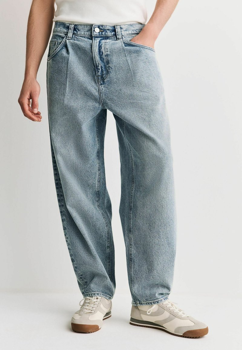 Homme portant un jean bleu clair ample, une chemise blanche rentrée et des baskets beiges avec des semelles marron, debout avec une main dans la poche.