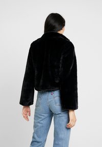 Chaqueta de piel sintética negra cortada con un cuello chal y mangas largas, llevada sobre jeans azul de cintura alta, que muestra una textura suave.