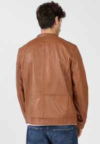 Braune Lederjacke mit glatter Textur, mit Nahtdetails und einem schlichten Kragen. Darüber wird ein weißes Shirt getragen, kombiniert mit Denim.