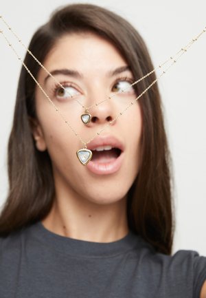 Joven con cabello largo y castaño, vistiendo una camisa oscura, mira dos collares de oro con colgantes de piedra preciosa en forma de triángulo que sostienen frente a su rostro.