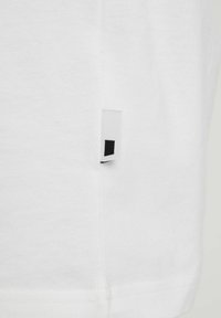 Weißes Baumwoll-T-Shirt mit sichtbarem schwarzem Quadrat-Etikett an der Seitennaht. Es zeichnet sich durch eine glatte Textur und ein sauberes, schlichtes Design aus.