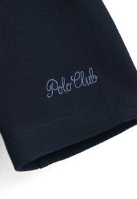 Granatowa bawełniana koszulka polo z teksturowanym materiałem; posiada haftowane w jasnoniebieskim kolorze logo "Polo Club" po lewej stronie blisko dołu.