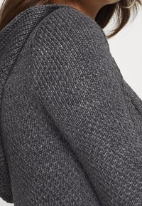 Grå stickad tröja med en strukturerad tyg, huva och intrikata sömmar längs sömmarna för extra detalj och struktur.