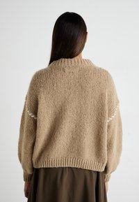 Beige strikkegenser med ribbestrikket krage og overdimensjonerte ermer. Har hvite sømdetaljer på skuldrene og en teksturert finish.