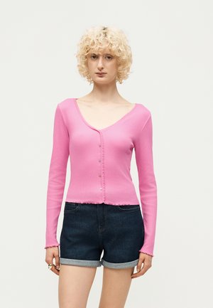 Osoba s krátkymi kučeravými blond vlasmi, oblečená v ružovom zapínanom tričku s dlhým rukávom a tmavých vyhrnutých rifľových šortkách na jednoduchom pozadí.