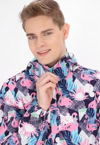 Jachetă colorată cu un fundal bleu marin, având flamingo roz, frunze tropicale și motive geometrice. Materialul pare ușor.