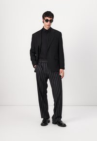 Black tailored blazer over a black shirt, paired with black and white striped trousers. Accessories include dark sunglasses and black shoes.