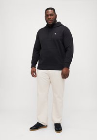 Black hooded sweatshirt with a front pocket, paired with white pants and black sneakers. Features white logo on the chest.