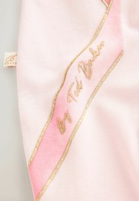 Heledad roosa kangas, kus on diagonaalne roosa lint, millel on kuldne ääris, ja mis kannab kätkestatud kuldsete tähtedega "by Ted Baker".