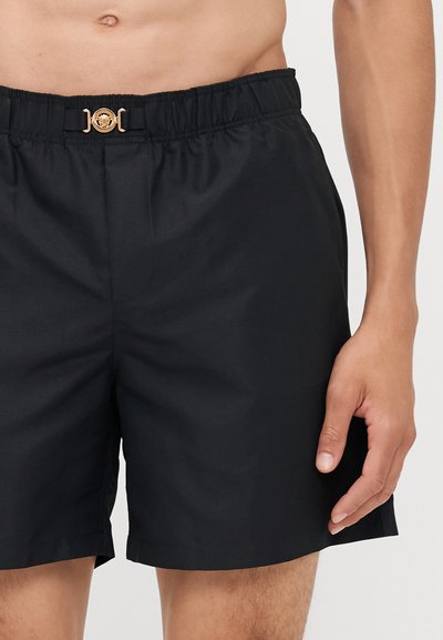 Shorts de bain noirs avec une taille élastique et un détail de boucle circulaire dorée à l'avant. Tissu lisse, coupe longueur genou.