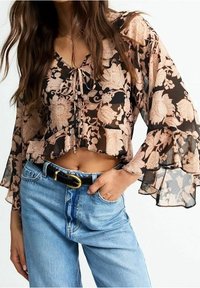 Blouse noire légèrement transparente avec des motifs floraux pêche, décolleté en V, détail à nouer et manches évasées, portée avec un jean bleu taille haute et une ceinture noire.