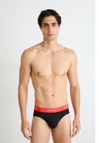 Fekete pamut alsónadrág piros elasztikus derékpánttal, amelyen a "Calvin Klein" logo látható, testhezálló kialakítás, egyszínű textúra és minimalista stílus.