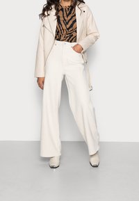 Pantaloni bianchi a gamba larga, un top aderente con stampa zebra marrone e nera, e una giacca da biker in pelle crema completano l'outfit.