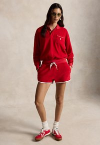 Kobieta ubrana w czerwoną koszulkę polo z długim rękawem, pasujące czerwone szorty z białym obszyciem, białe skarpetki, czerwone buty sportowe i zielone okulary przeciwsłoneczne, stojąca z rękami w kieszeniach.