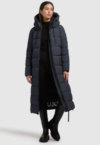 Lungo cappotto puffato blu navy con cappuccio, caratterizzato da un design trapuntato, chiusura frontale con zip e dettagli elasticizzati. Abbinato a stivali neri al ginocchio.