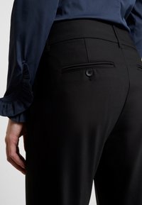 Pantalons noirs avec une texture lisse, dotés d'une poche arrière et de détails en bouton. Un chemisier bleu foncé à manches volantes est partiellement visible.