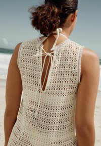 Cubierta de playa de punto en beige claro con patrón de tejido abierto. Presenta un cierre con lazo en el cuello y bordes festoneados en las mangas.
