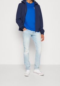 Granatowa kurtka na zamek błyskawiczny na niebieskiej koszulce, w zestawie z jasnoniebieskimi dżinsami i białymi trampkami. Codzienny strój o luźnym kroju.