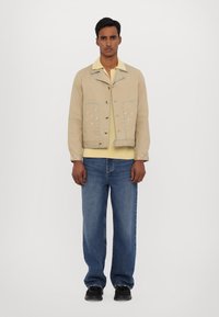 Giacca beige con cuciture decorative, tasche frontali e bottoni. Camicia gialla sotto, abbinata a jeans di denim azzurri e scarpe nere.